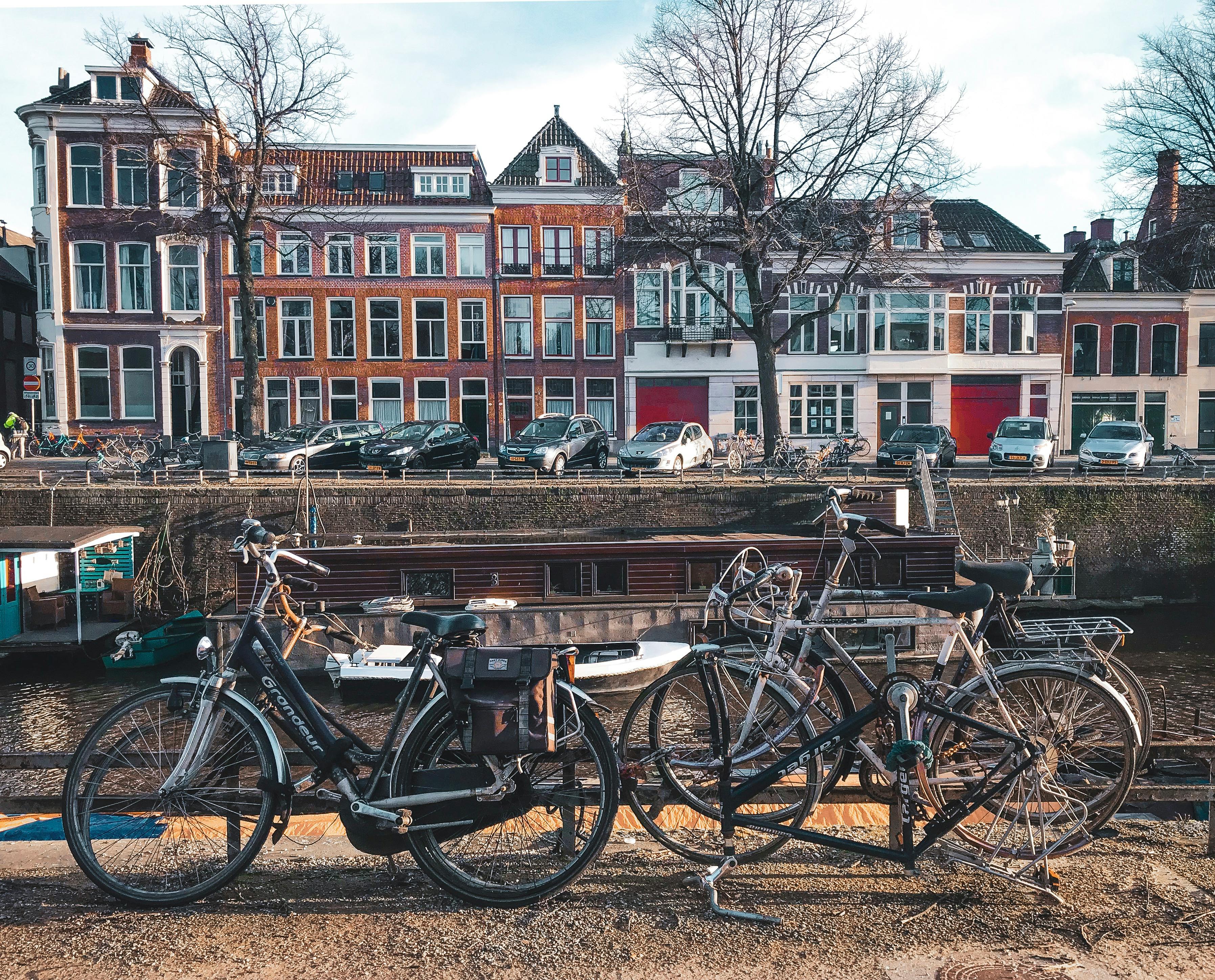 A view of Groningen. A canal, with bikes lined up before the water. Houses face us across the water, with cars parked up.