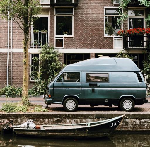 A blue van parked in front of a brown brick stone apartment building. A boat bobs by on the canal in front.