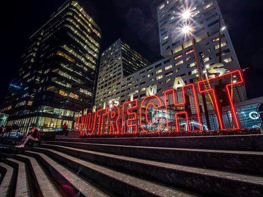 Steps in central Utrecht at night, with large neon letters reading 'In Utrecht' in the center. Behind are tall apartment buildings.